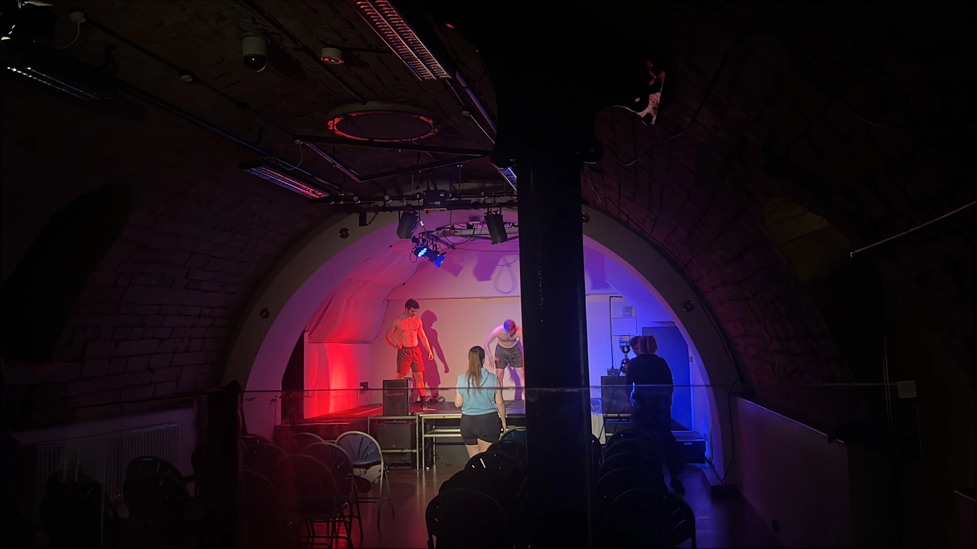 A stage photo from the back of an empty theatre. Three people stand onstage. The stage is lit with the left half in red and the right half in blue.
